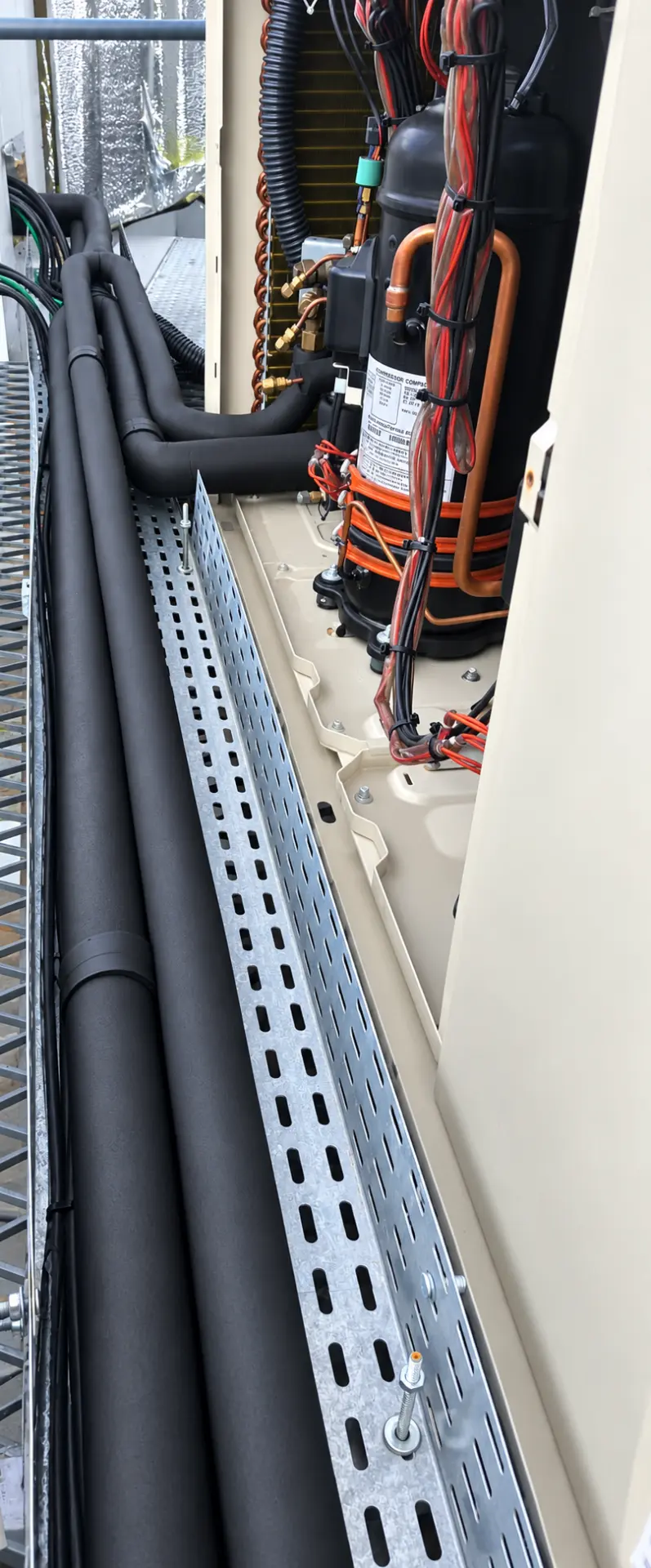 Detalhe interno de unidade de climatização com compressor, tubulação isolada e bandeja de cabos